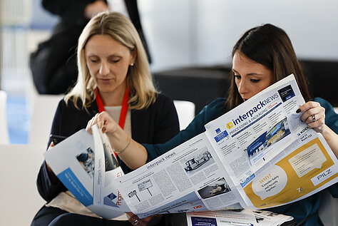 Two women seated and reading an issue of the ‘interpack news’ newspaper, focusing on event-related content.