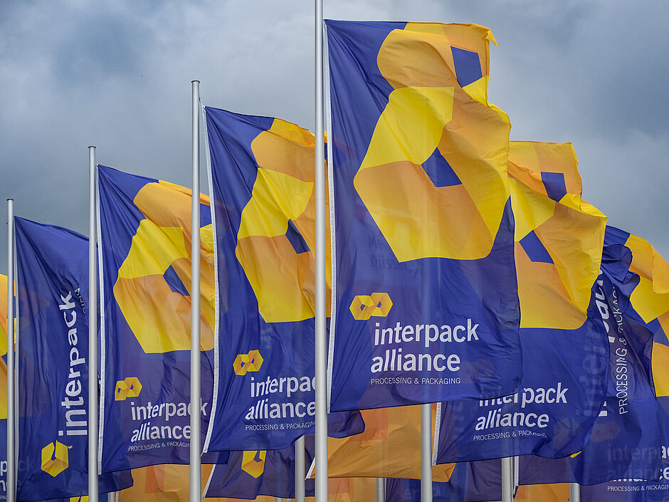 Several blue flags waving, featuring the yellow-orange logo and the text ‘interpack alliance – Processing & Packaging’ against a cloudy sky.