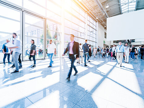 A bright, sunlit exhibition hall with many people walking through the spacious interior with large windows casting shadows on the floor.